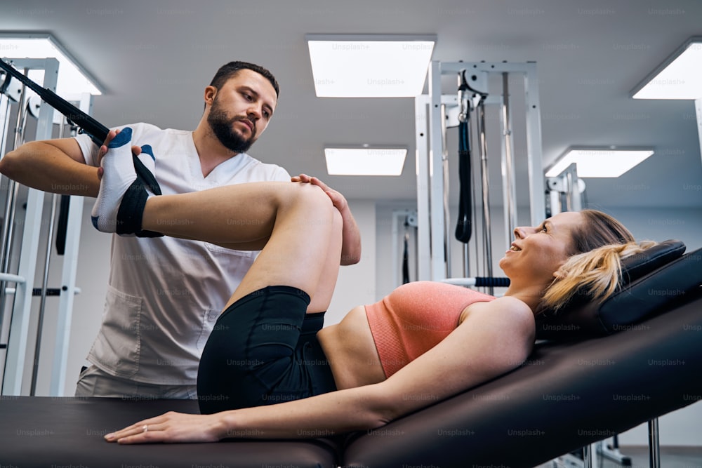 Massagem e treinamento de força de pernas de mulheres jovens em máquina elástica para reabilitar lesões esportivas. Fisioterapeuta masculino trata coluna vertebral e costas em tratamento fisioterapêutico