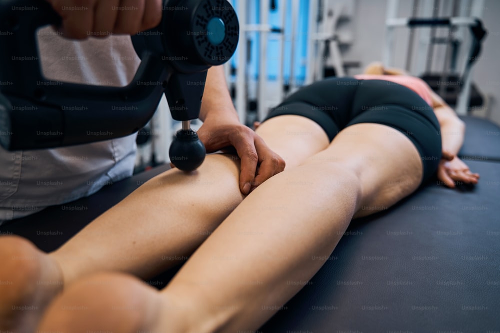 Massagem de martelo de percussão close up para mulher em consultório terapêutico de centros de reabilitação. Homem massagista dando massagem profunda aos músculos das pernas e nádegas para regeneração. Lesão esportiva