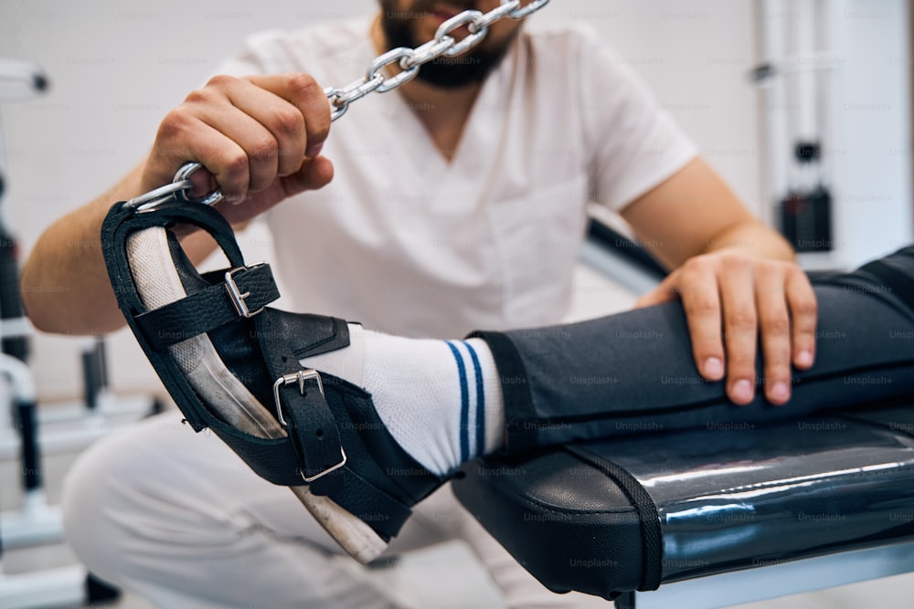 Nahaufnahme des Physiotherapeuten, der eine junge Frau mit Rehabilitationsschuhen zu Fuß auf einem elastischen Kraftsimulator trainiert. Erholung mit Reha-Ausrüstung. Patientin bei Beintherapie nach Sportverletzung