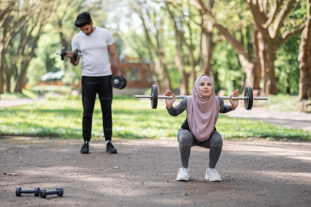 Fitness-Frau in Sportkleidung und Hijab hockt mit Langhantel, während ein starker muslimischer Mann die Arme mit Hanteln trainiert. Aktiver Lebensstil der jungen Familie.