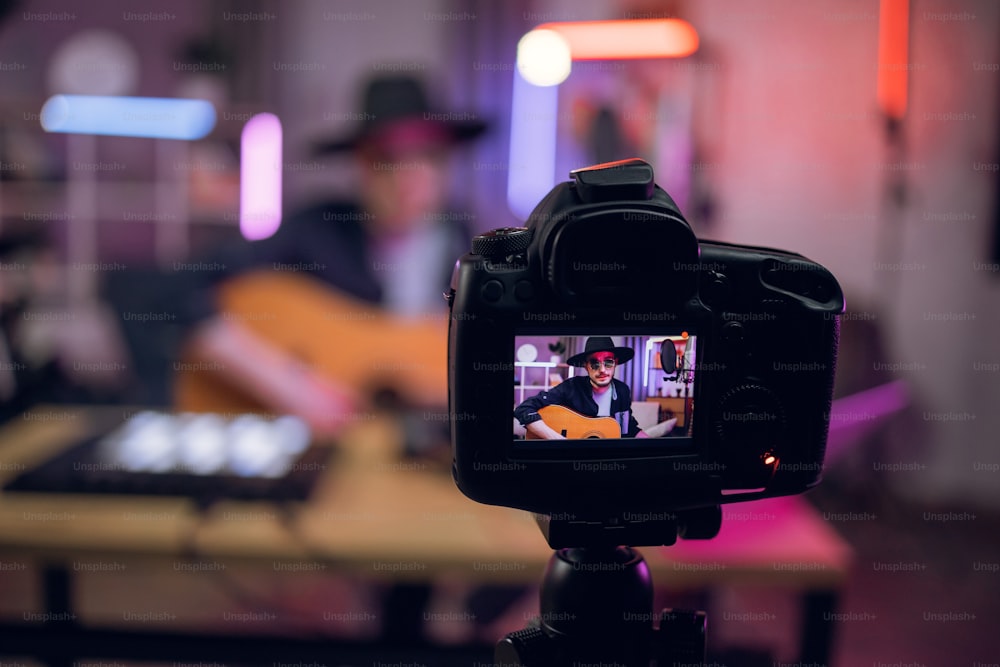 Blur background of male artist in hat and sunglasses playing guitar and recording broadcast. Focus in digital camera fixed on tripod. Music concept.
