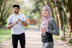 Beautiful woman in sport clothes and hijab holding bottle of water while standing at green park. Blur background of handsome muslim guy refreshing and relaxing after run.