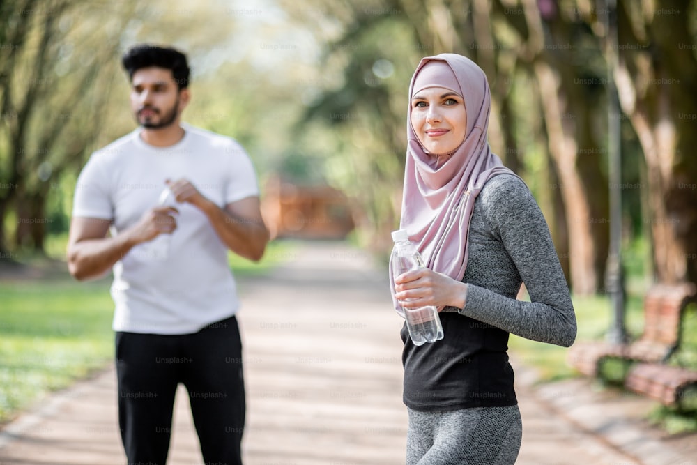 Hermosa mujer con ropa deportiva y hiyab sosteniendo una botella de agua mientras está de pie en el parque verde. Fondo borroso de chico musulmán guapo que se refresca y se relaja después de correr.