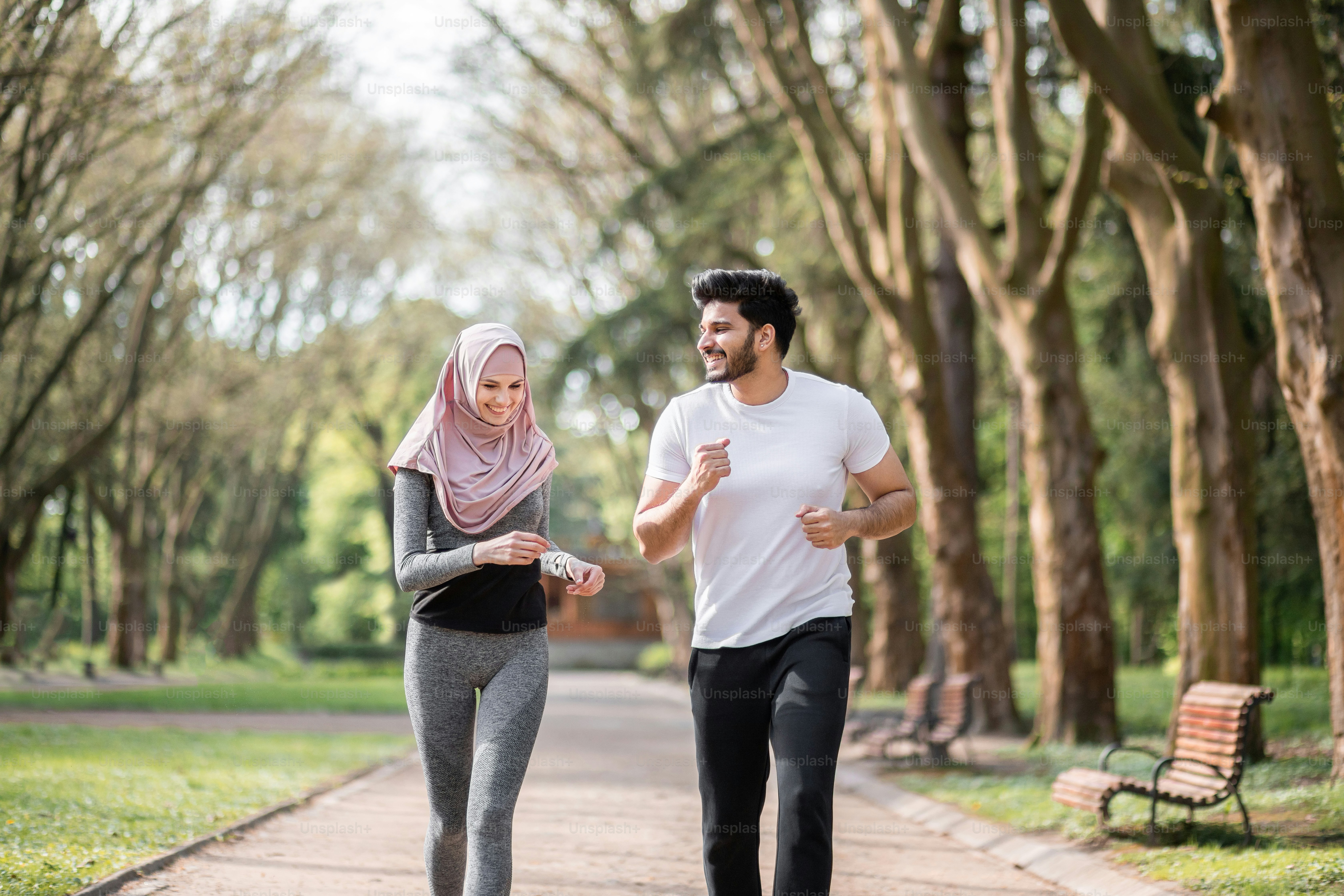 Jóvenes positivos con ropa deportiva corriendo juntos en el parque verde de la mañana. Esposo musulmán guapo y esposa encantadora en hiyab entrenando activamente al aire libre.