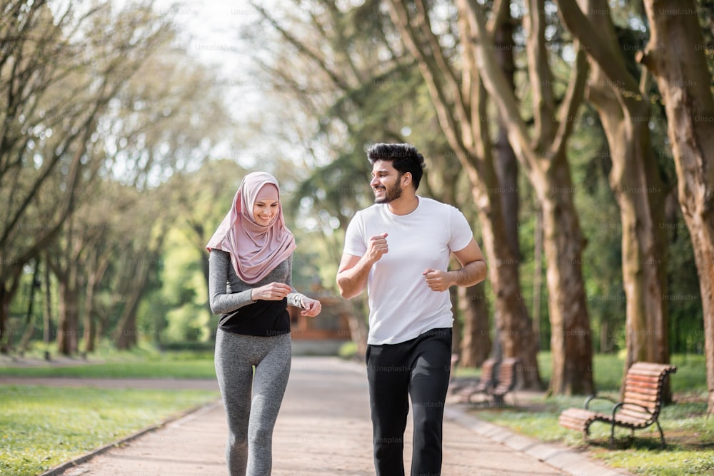 Positive junge Menschen in Sportkleidung laufen gemeinsam im morgendlichen grünen Park. Hübscher muslimischer Ehemann und charmante Frau im Hijab, die aktiv im Freien trainieren.