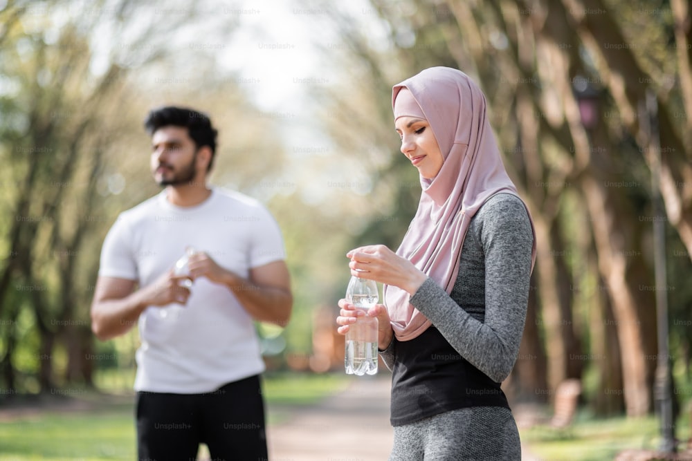 Young woman sport clothes and hijab holding bottle with water while standing at green park. Blur background of her muslim handsome husband that looking aside.