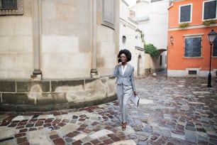 Successful african woman in business suit using modern smartphone for conversation outdoors. Young business lady walking on city street and carrying clipboard.