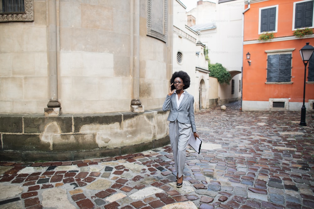 Successful african woman in business suit using modern smartphone for conversation outdoors. Young business lady walking on city street and carrying clipboard.