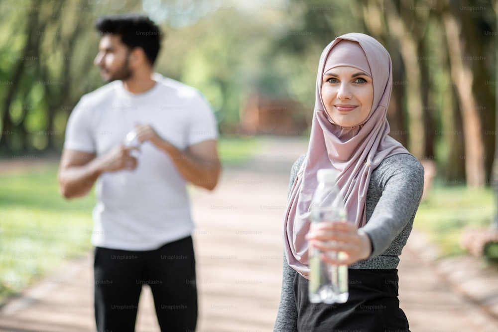 Hübsche Frau im Hijab, die Wasser der Flasche hält, während ein muslimischer Typ dahinter steht und zur Seite schaut. Junge Familie in Activewear erneuert den Wasserhaushalt nach dem Training im Freien.
