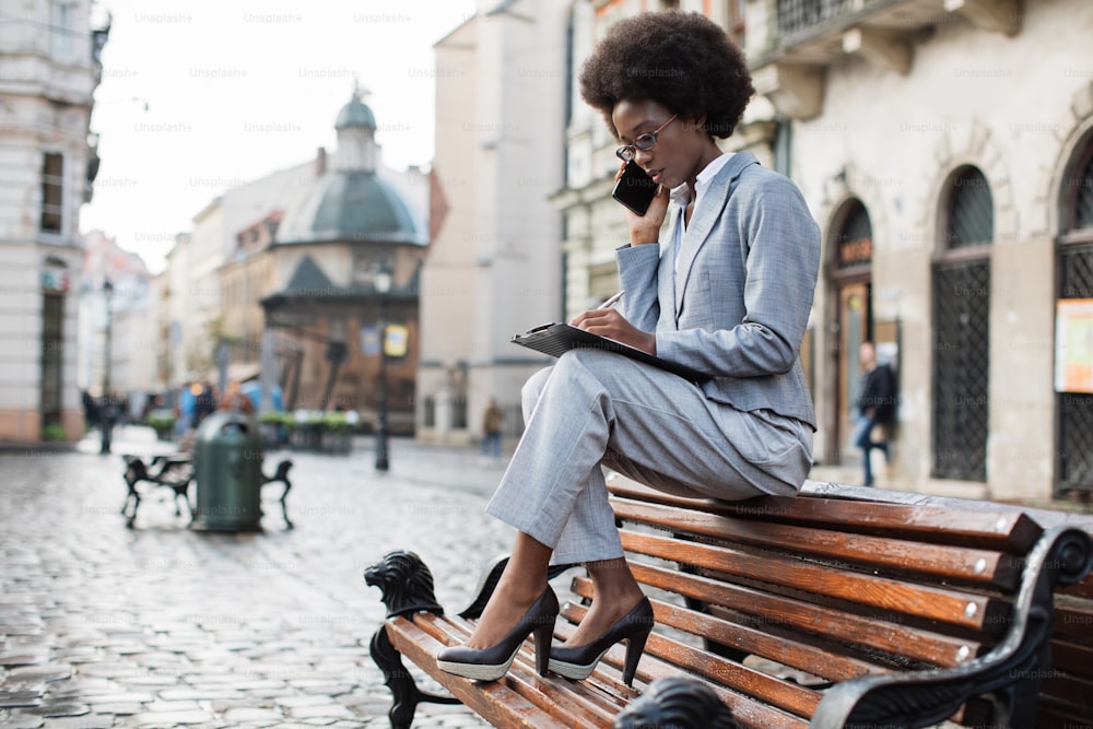 Attractive african woman in business suit sitting on wooden bench and talking on mobile. Serious lady taking notes on clipboard during conversation on fresh air.