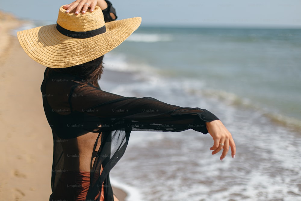 Stylish carefree woman holding hat and relaxing on sunny beach at sea, close up. Summer vacation. Young fashionable female in straw hat and light shirt enjoying holiday on tropical island