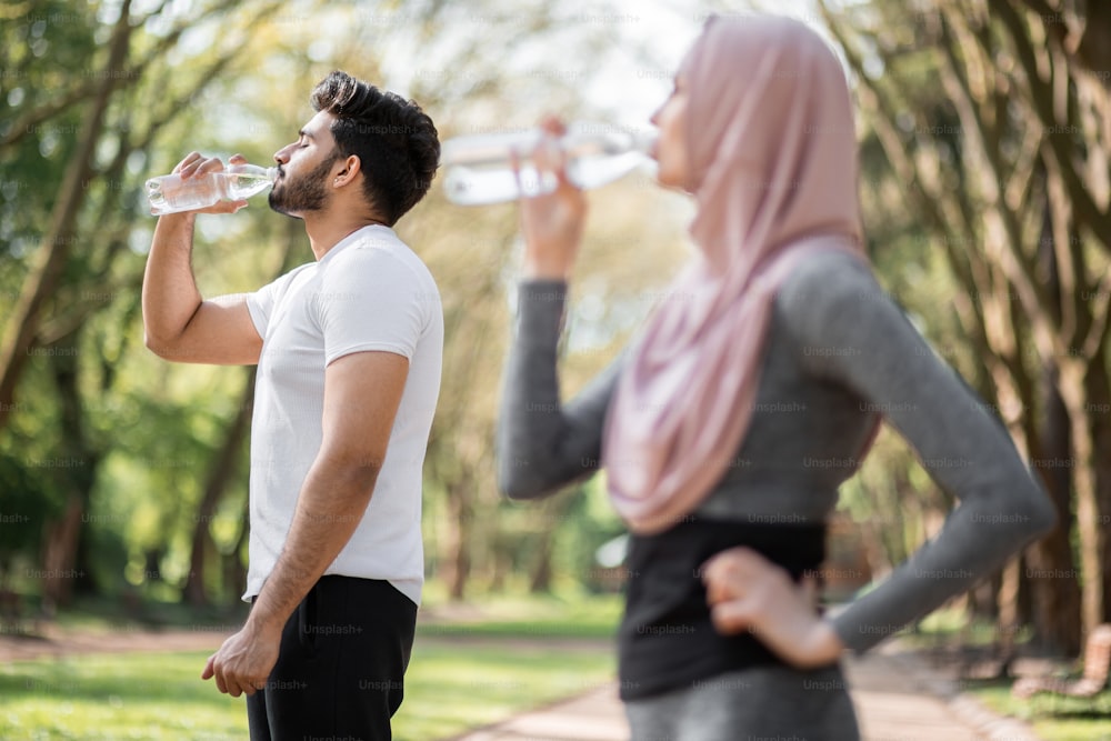 Active muslim couple in sport outfit standing together at green park and drinking fresh water. Blur foreground of woman in hijab. Outdoors activity and recreation concept.