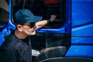 Professional truck driver with hat and sunglasses confidently standing in front of big and modern truck. Bright sunny day. People and transportation concept.