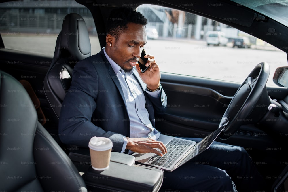 Uomo d'affari serio in abito formale che parla sul cellulare e digita sul laptop mentre è seduto in un'auto elettrica di lusso. Uomo africano che risolve momenti di lavoro a distanza.