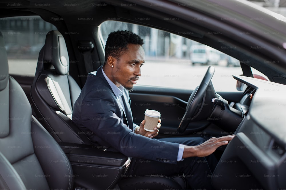 Side view of focused african man sitting on driver's seat with cup of coffee and using dashboard for navigation. Businessman in formal wear taking stop for checking direction.