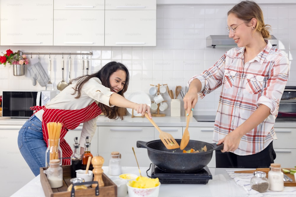 Cute joyful couple cooking together and adding spice to meal, laughing and spending time together in the kitchen, lesbian couple in apron, smiling and dancing together behind a wooden table with frying pan
