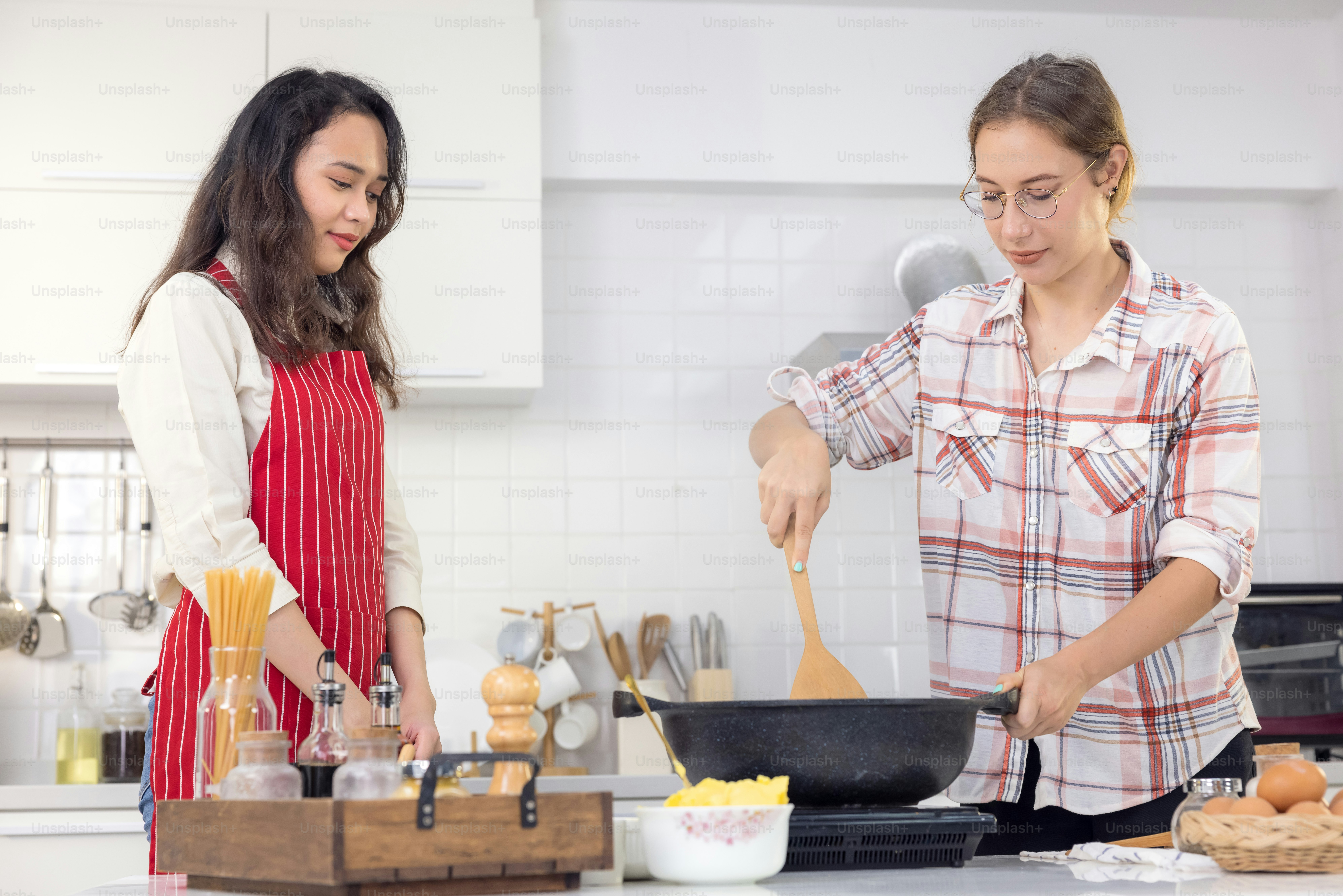 Couple joyeux mignon cuisinant ensemble et ajoutant des épices au repas, riant et passant du temps ensemble dans la cuisine, couple de lesbiennes en tablier, souriant et dansant ensemble derrière une table en bois avec poêle à frire
