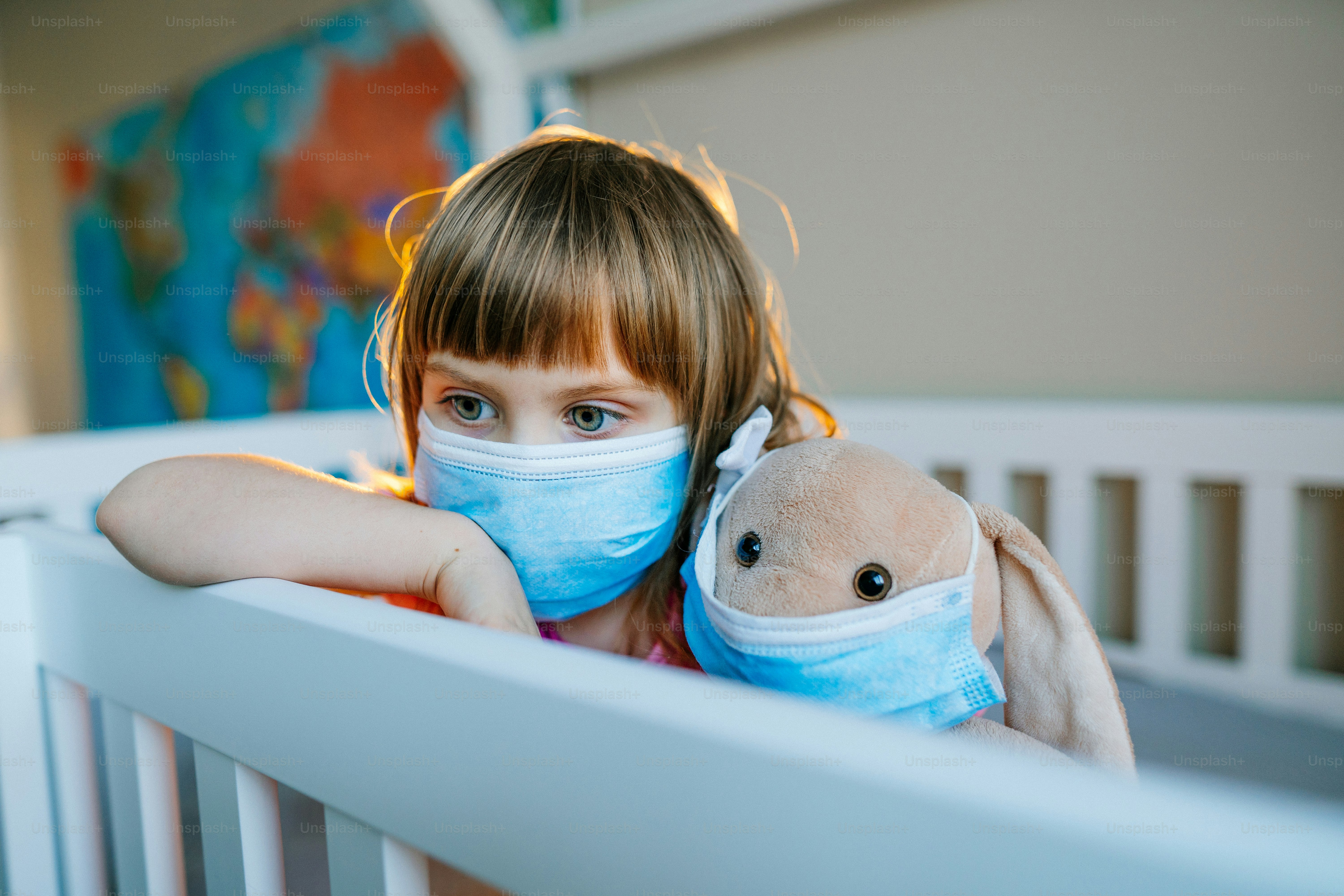 Kleines 4-jähriges Mädchen mit Schutzmaske spielt mit ihrem Plüschtierhasen, das auf dem Bett im Kinderzimmer sitzt. Neue Normalität nach pandemischem COVID-19-Konzept.