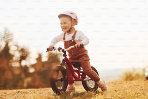 Glückliches kleines Mädchen mit Schutzhut, das an sonnigen Tagen in der Nähe des Waldes mit dem Fahrrad im Freien fährt.