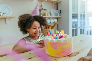 キッチンでの誕生日パーティー中に楽しい時間を過ごしているキャンドル付きのバースデーケーキを見ている幸せな女の子。女の子の顔に選択的に焦点を合わせます。