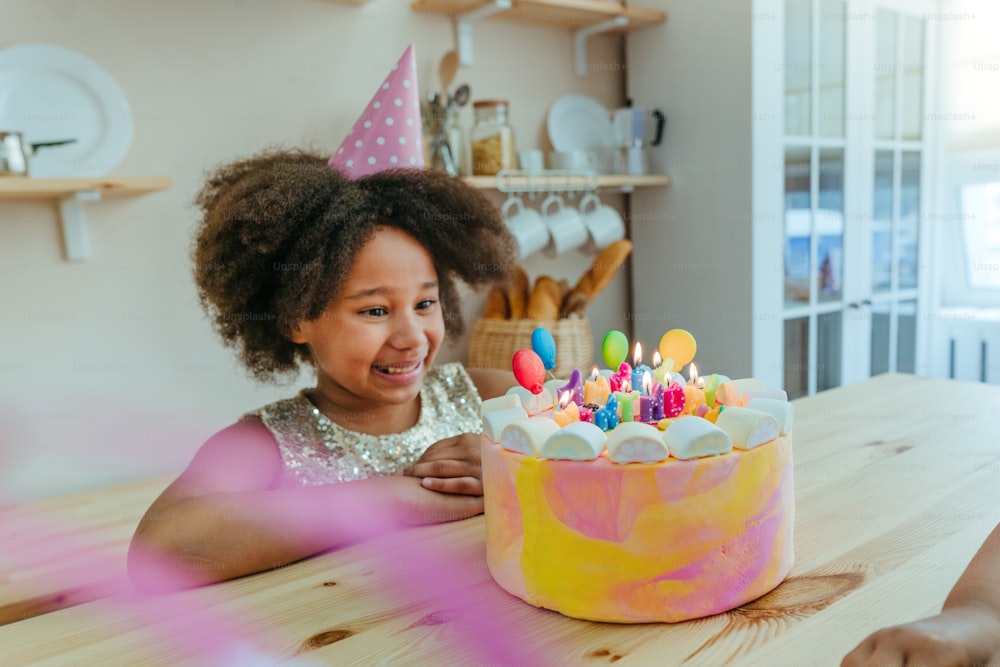 Happy girl looking at the birthday cake with candles having fun time during birthday party in the kitchen. Selective focus on the girl's face.