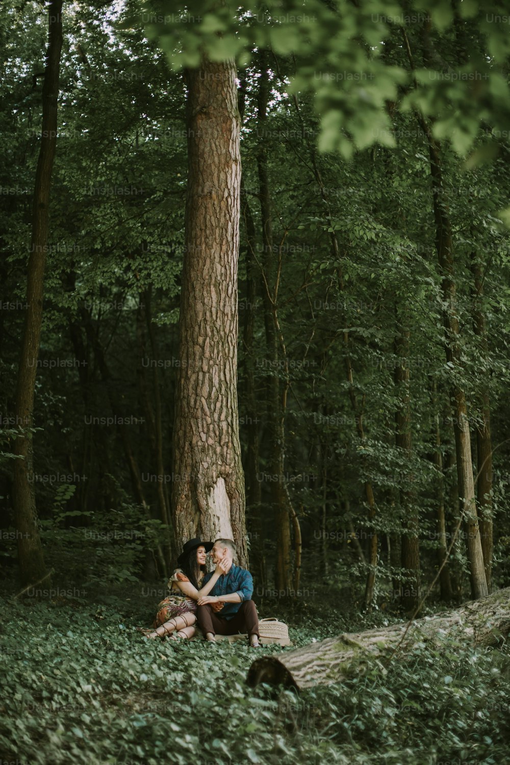 Outdoor full length shot of happy young family, man and woman, enjoying walk in the forest, sitting under the big tree, hugging and kissing. Pregnant couple sitting under the tree in forest