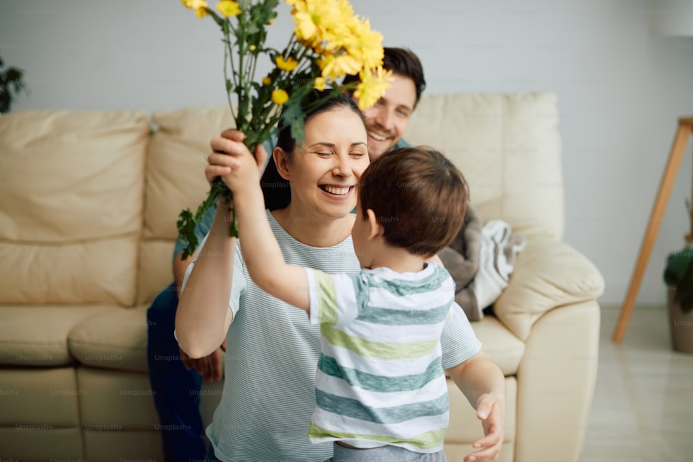 Small boy surprising his mother with bouquet of flowers at home. Focus is on mother.