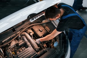 Under the hood. Man in work uniform repairs white automobile indoors. Conception of automobile service.