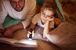 Lendo livro e usando lanterna. Pai e seu jovem jovem passando tempo juntos dentro de casa perto da tenda.