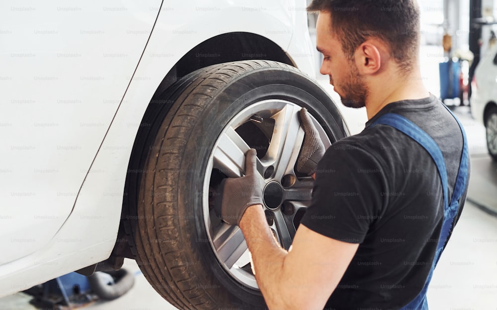 Homem no uniforme de trabalho mudando a roda do carro dentro de casa. Concepção de serviço automobilístico.