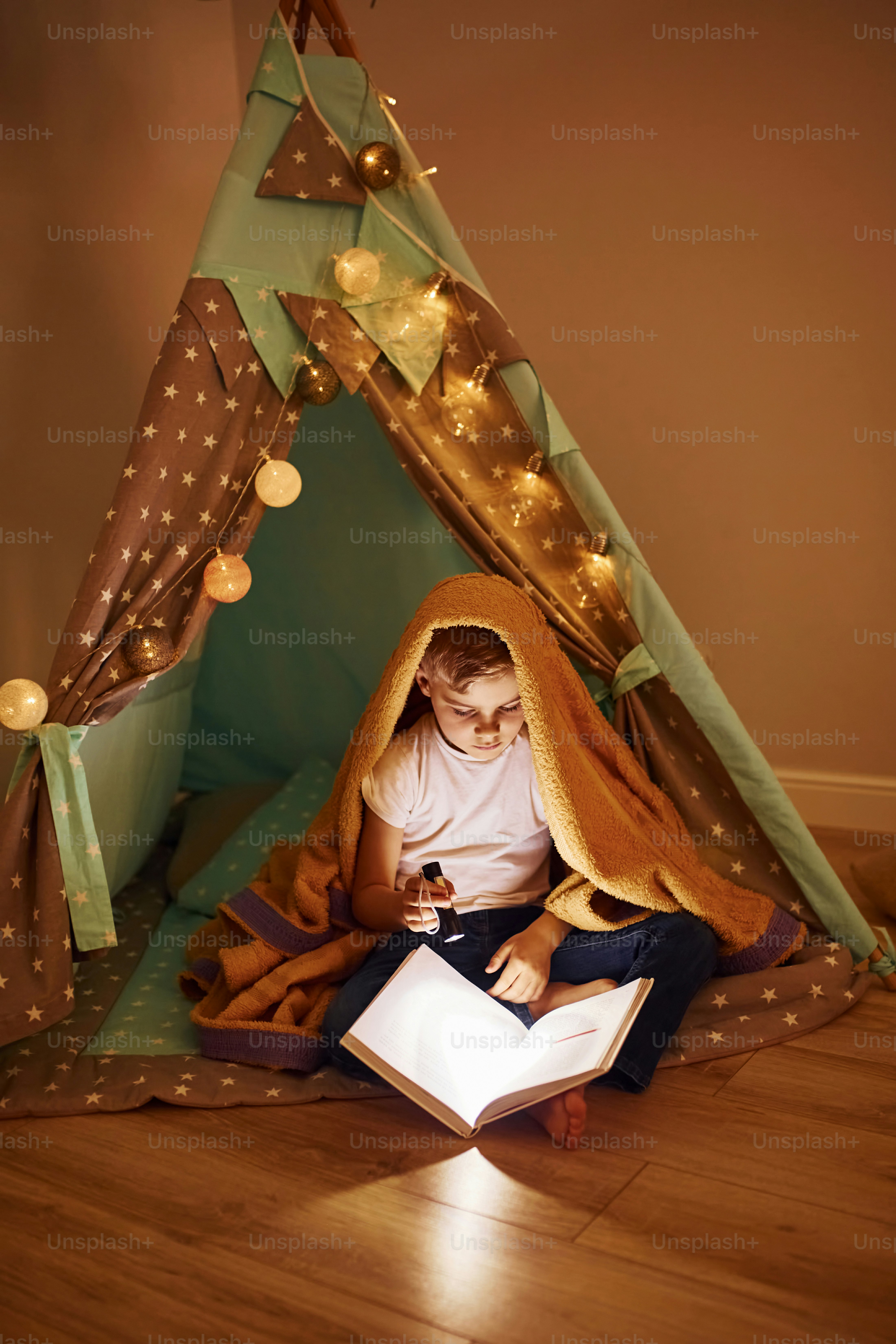 Reading book and using flashlight. Young boy in casual clothes lying down near tent at evening time.