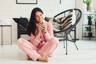Cheerful young woman in pajamas sitting on the floor indoors at daytime.