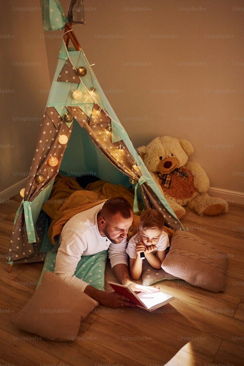 Buch lesen und Taschenlampe benutzen. Vater und seine jungen Jungen verbringen Zeit zusammen drinnen in der Nähe des Zeltes.