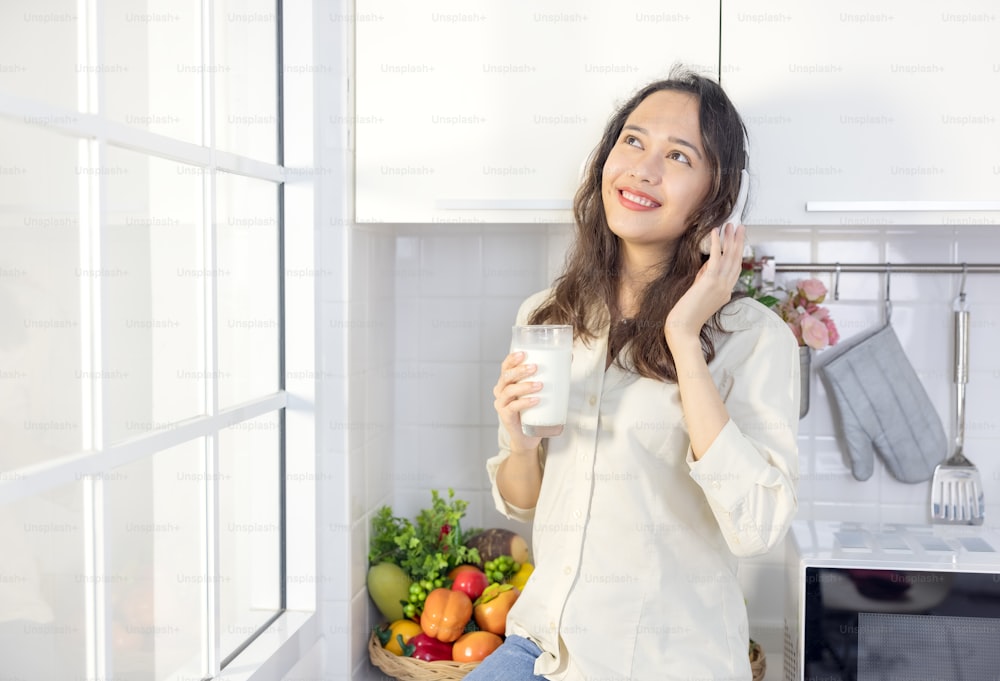 In a light modern kitchen, a beautiful woman drinks milk while listening to music. Before breakfast, she's having a good time and smiling.
