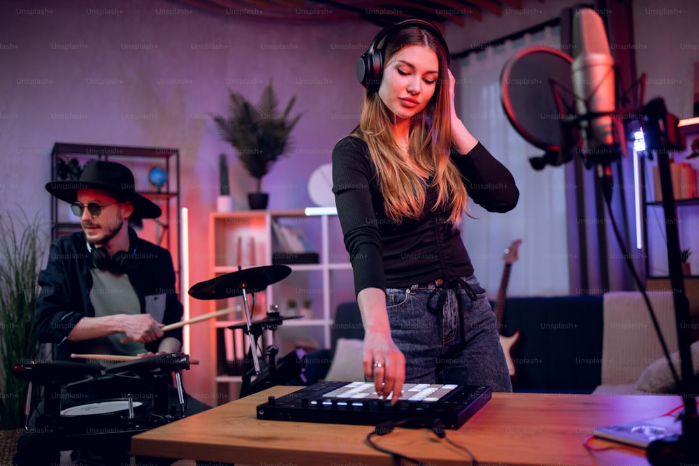 Adorabile giovane donna in cuffie che lavora sul mixer mentre l'uomo elegante in cappello e occhiali da sole suona la batteria elettronica. Processo creativo in studio di registrazione.