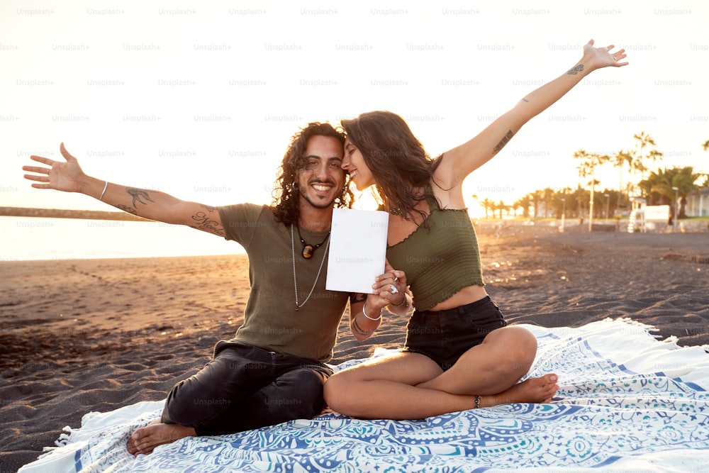 Hermosa pareja feliz en una cita, mostrando una tarjeta blanca vacía en la playa tropical. Vibraciones de vacaciones de verano. Hora de puesta de sol.