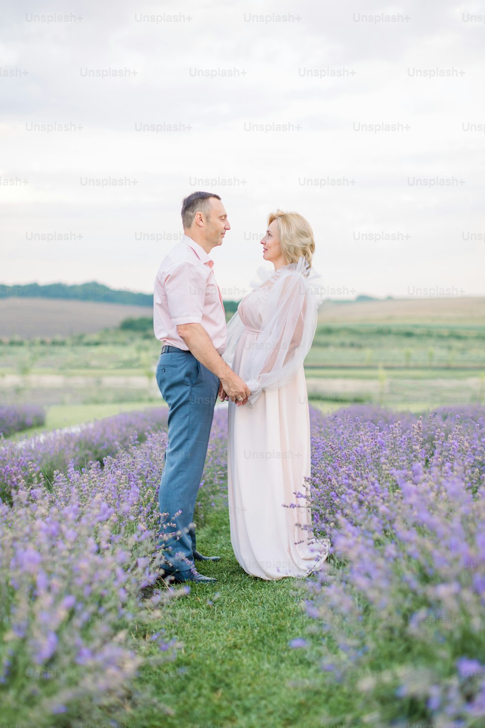Family portrait of middle aged handsome man and pretty woman, standing outdoors on lavender field, holding hands and looking each other. Wedding anniversary concept. Love through the years