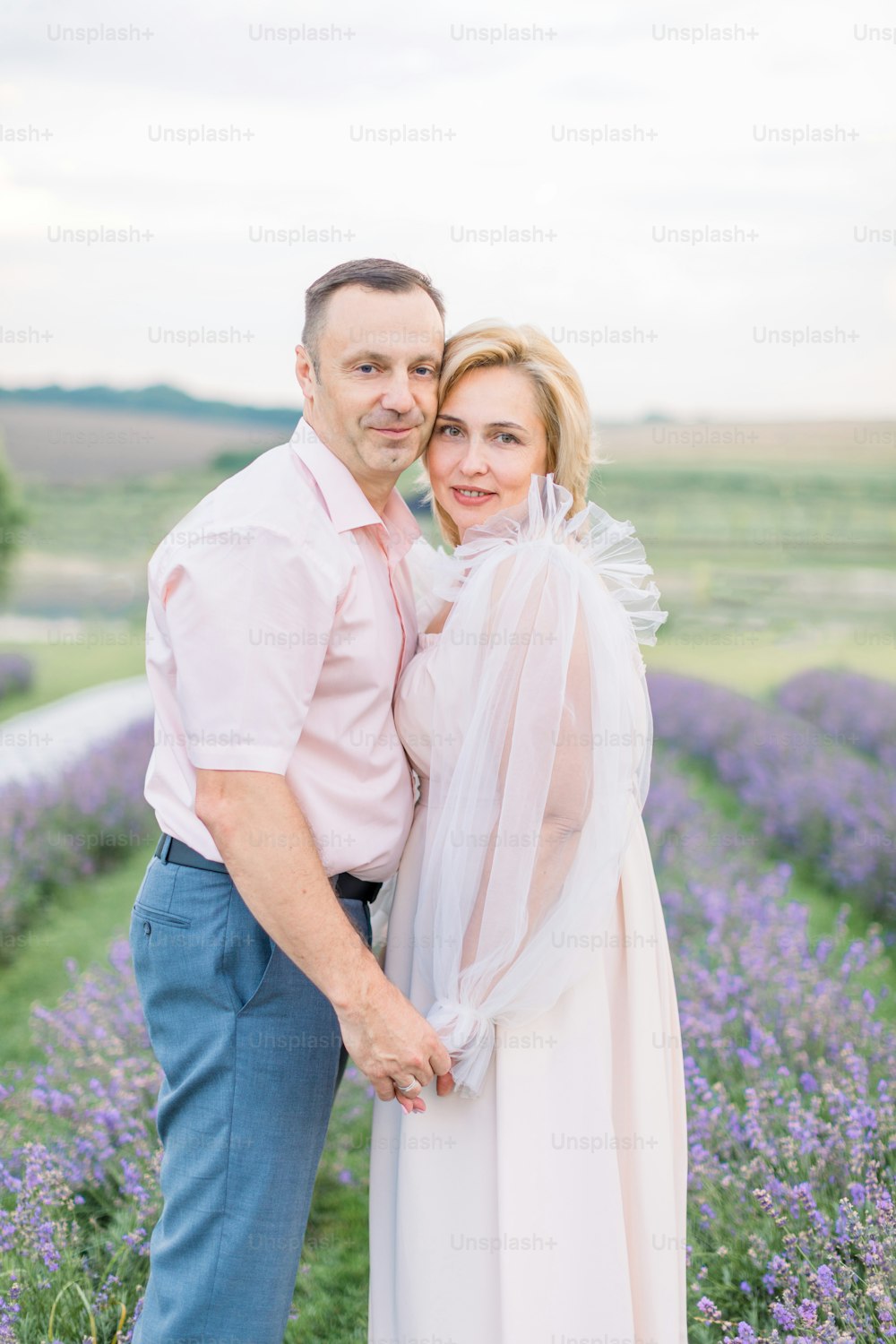 Front close up view of lovely mature couple at the lavender field, holding hands and hugging. Photo of caring man and woman walking outdoor in lavender field