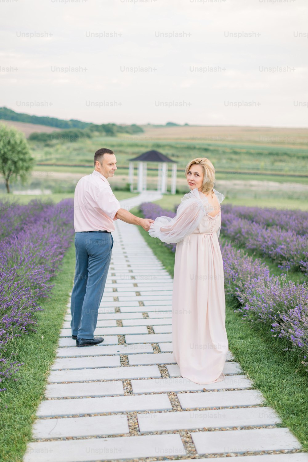 Outdoor shot of happy romantic mature couple in love walking on pathway through flowering lavender field, holding hands and enjoying moments together