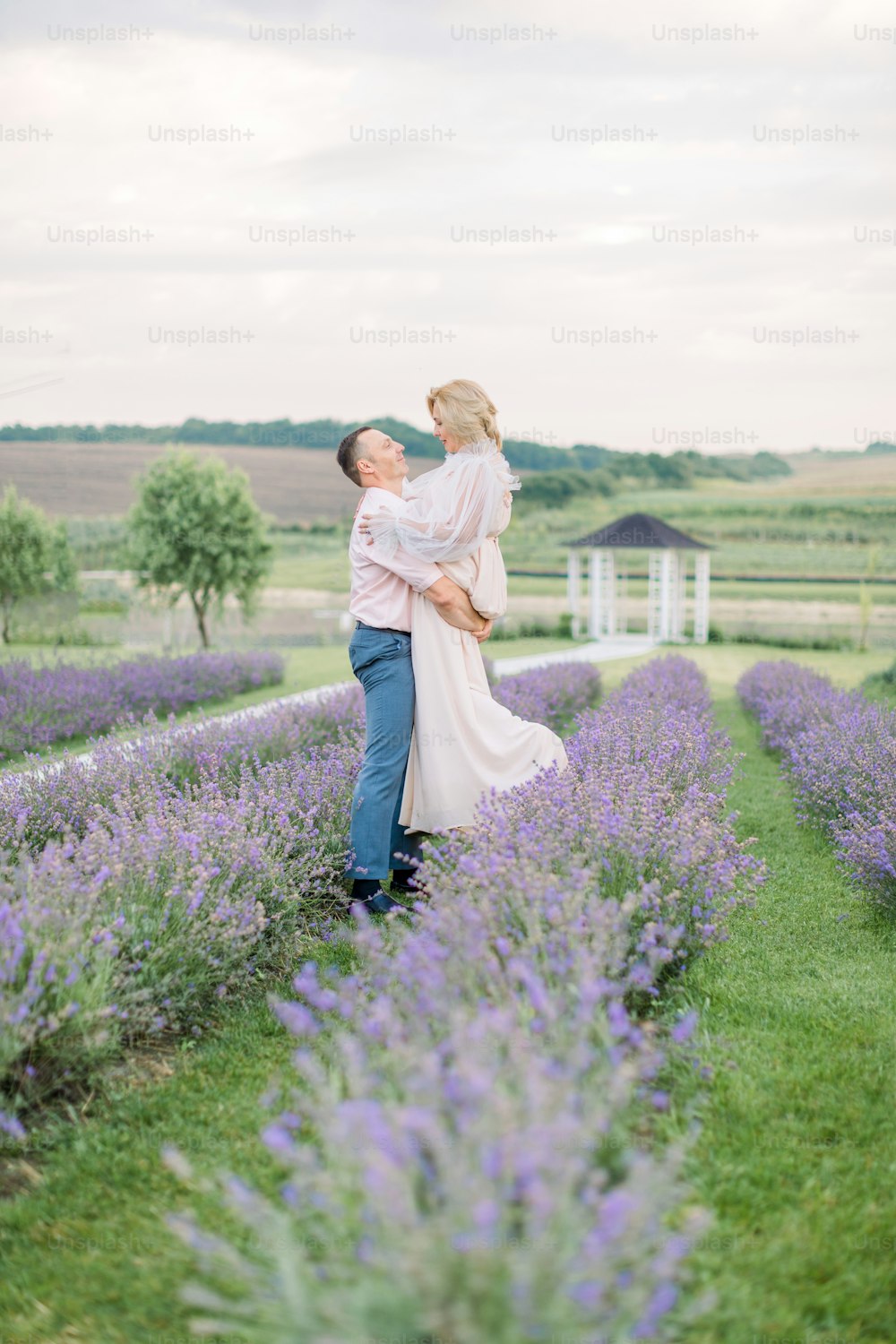 Happy middle aged lovely couple in purple lavender flower field, have romantic time and wedding anniversary celebration. Man holds his woman on hands and dancing