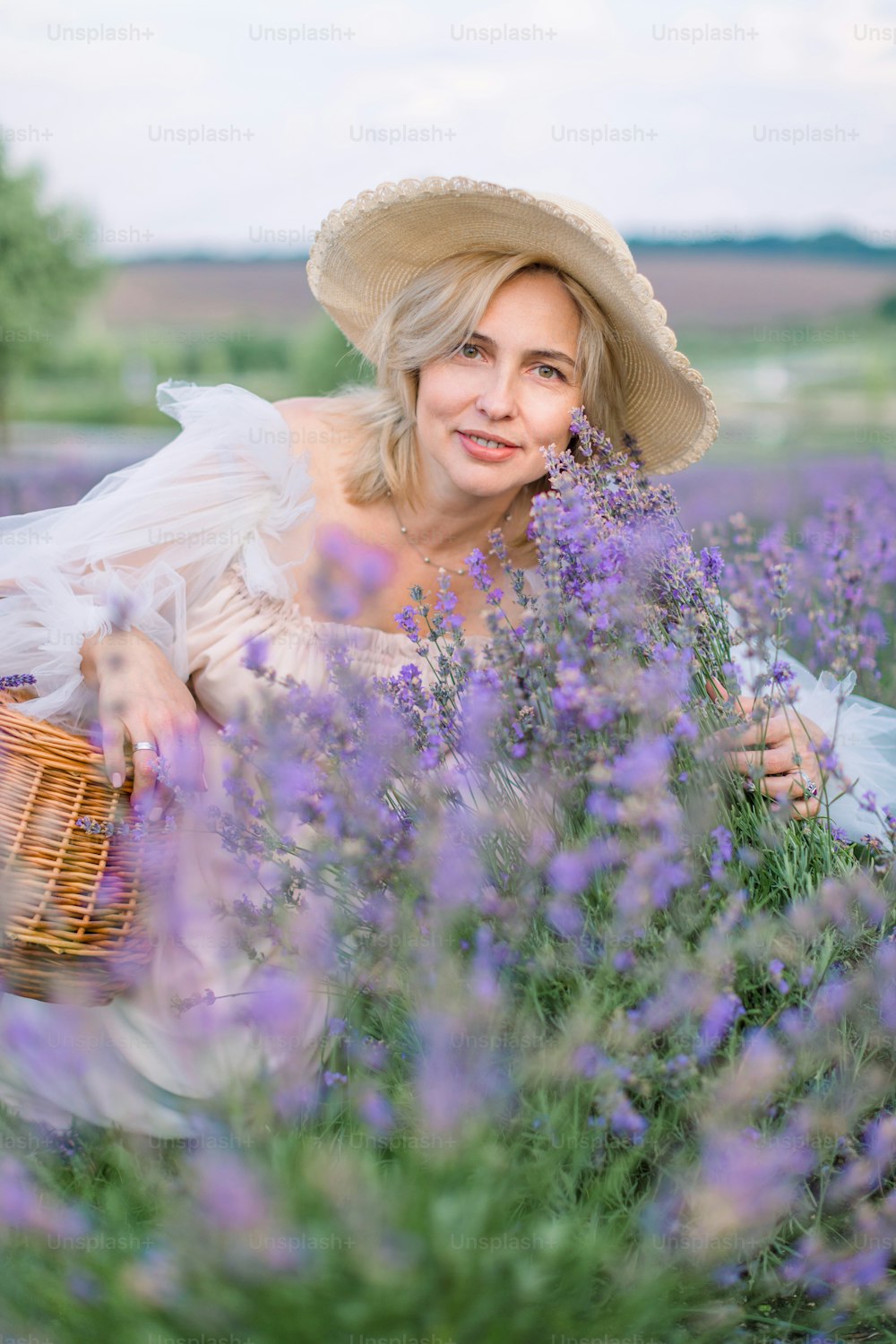Beautiful mature blond lady in lavender field. Smiling attractive middle aged woman in white dress and hat posing in a lavender field with small wicker basket