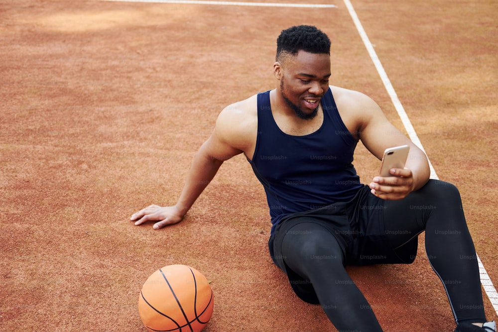 Utilizzo del telefono. L'uomo afroamericano gioca a basket sul campo all'aperto.