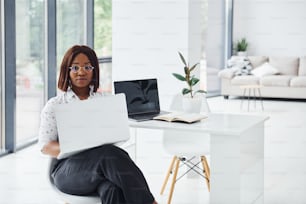 Joven afroamericana con ropa formal está en la oficina con una computadora portátil.