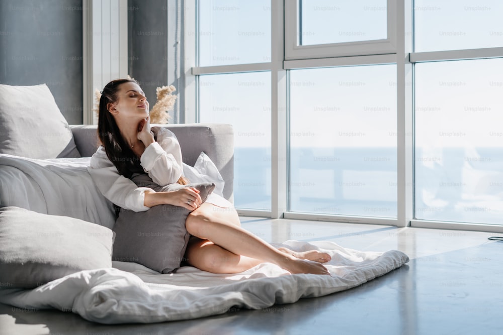 Young happy woman enjoying new day and sunshine coming through big panoramic window while sitting on floor in morning after waking up, keeping eyes closed and dreaming about something