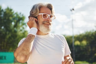 Listening to the music. Senior modern stylish man outdoors on the sportive field at daytime.