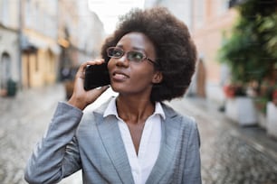 Attractive afro business woman in eyeglasses and suit talking on mobile and looking aside while standing on city street. Concept of people, work and conversation.