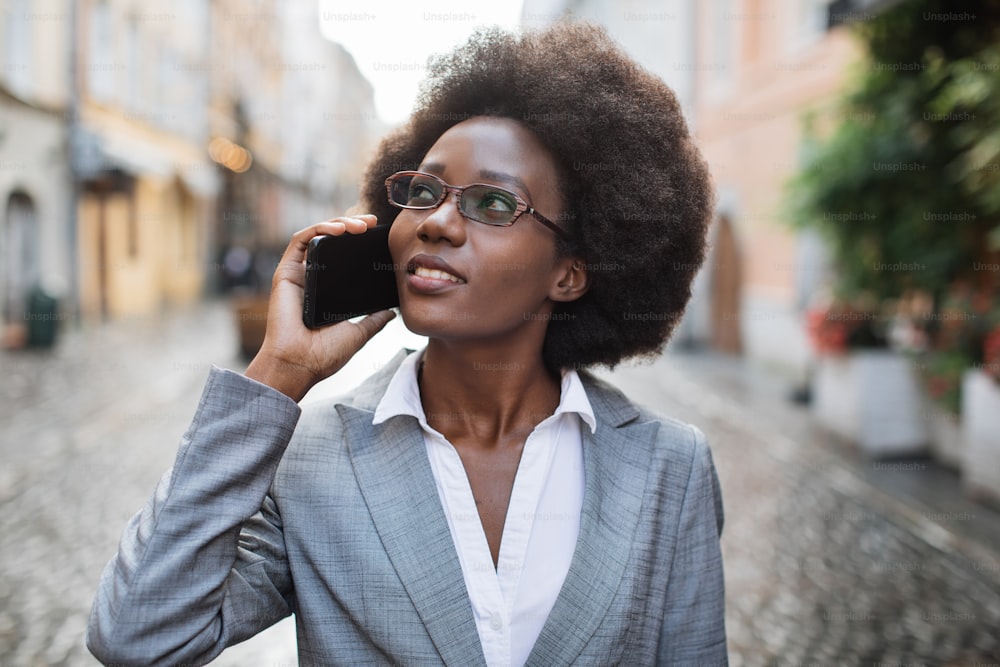 眼鏡とスーツを着た魅力的なアフロのビジネスウーマンが携帯電話で話し、街の通りに立って脇を見ています。人、仕事、会話のコンセプト。
