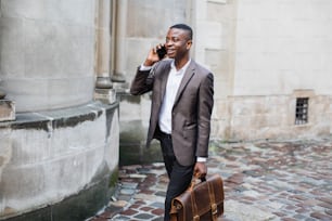 Lächelnder Afro-Amerikaner im Business-Anzug, der auf dem Handy spricht, während er mit einer Lederaktentasche in der Hand auf der Straße geht. Konzept von Kommunikation und Technologie.
