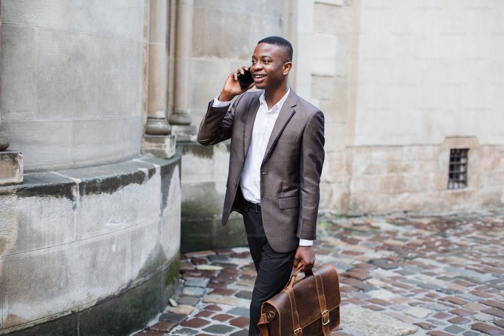 Smiling afro american man in business suit talking on mobile while walking on street with leather briefcase in hand. Concept of communication and technology.