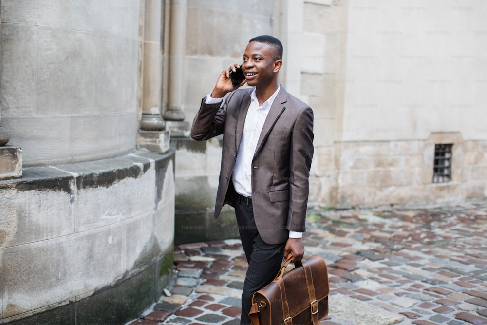 革のブリーフケースを手に通りを歩きながら、携帯電話で話しているビジネススーツを着た笑顔のアフロアメリカ人男性。コミュニケーションとテクノロジーの概念。